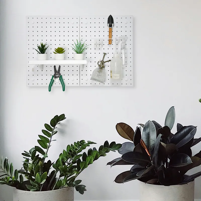 White modular pegboard system mounted on a white wall above indoor plants, organized with gardening tools and small succulents.